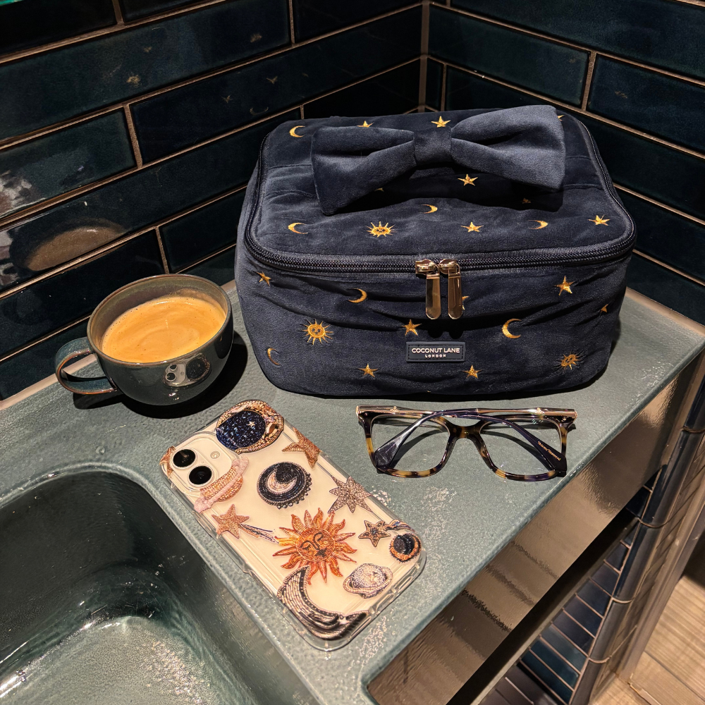 Clear phone case with celestial pattern next to a makeup bag, glasses, and mug on blue tiles.