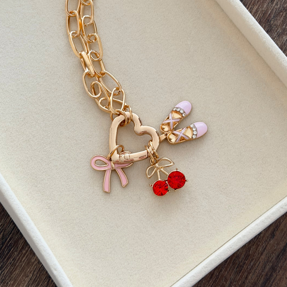 A gold chain necklace with a heart pendant, crescent moon, flower, and star charms. "THE CHARM STUDIO" text appears in black in the lower right corner on a white background.