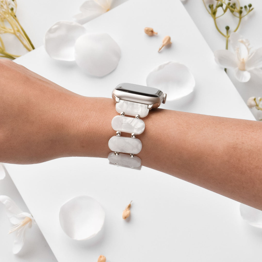A person’s arm with a smartwatch on their wrist featuring a white, semi-transparent band composed of large, smooth, rectangular links. The background is decorated with white petals and small, delicate flowers.