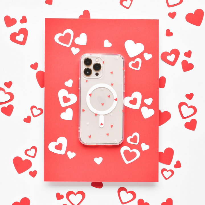A smartphone with a high-quality Coconut Lane Clear MagSafe® Phone Case - Cute Hearts, adorned with tiny red hearts, is placed on a red card. The setup is surrounded by scattered red and white heart-shaped cutouts, creating a romantic and festive theme.