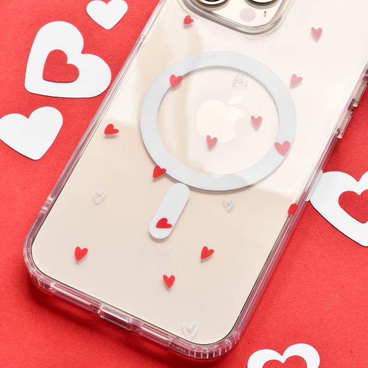 A high-quality protection smartphone with a Clear MagSafe® Phone Case - Cute Hearts by Coconut Lane is resting on a red surface decorated with white and red heart shapes. The phone case has a circular design and tiny red hearts, allowing the Apple logo on the back of the phone to be visible.