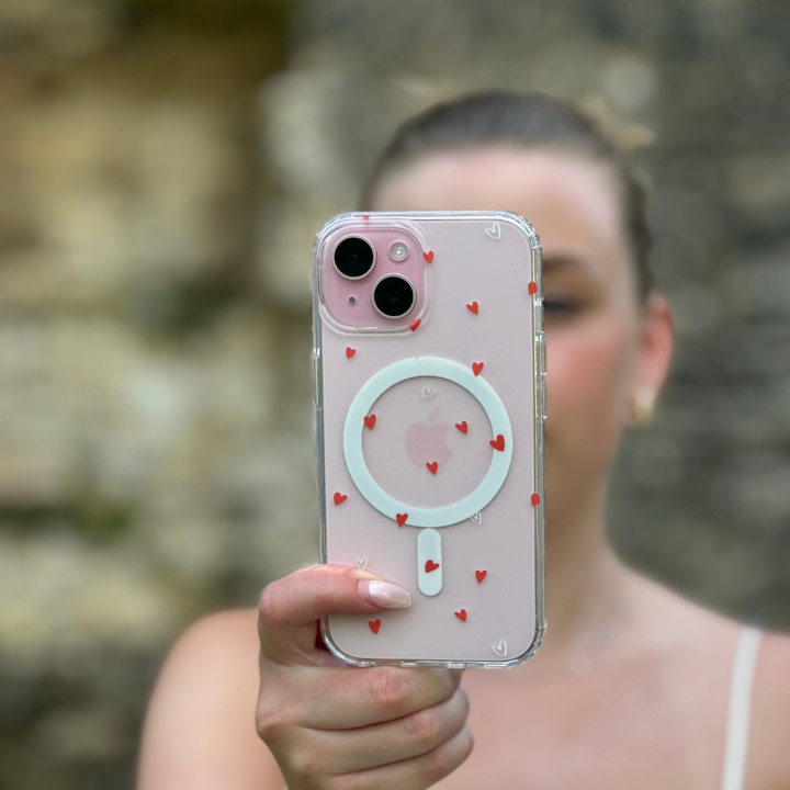 A person holds a pink iPhone covered with the Clear MagSafe® Phone Case - Cute Hearts by Coconut Lane. Small red hearts are scattered across the case, providing both high-quality protection and an aesthetic touch. The MagSafe® technology circle is visible through the case. The background is blurry, and the person is slightly out of focus, bringing attention to the phone.