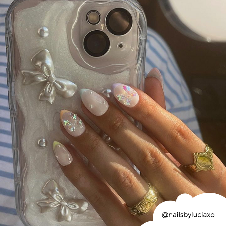 Close-up of a hand with manicured nails over the Pearls & Bows Phone Case by Coconut Lane, adorned with 3D bows and gems. The almond-shaped nails are covered in glossy, light pink and white polish, featuring holographic nail art. The hand is adorned with multiple gold rings. Text: @nailsbyluciaxo.