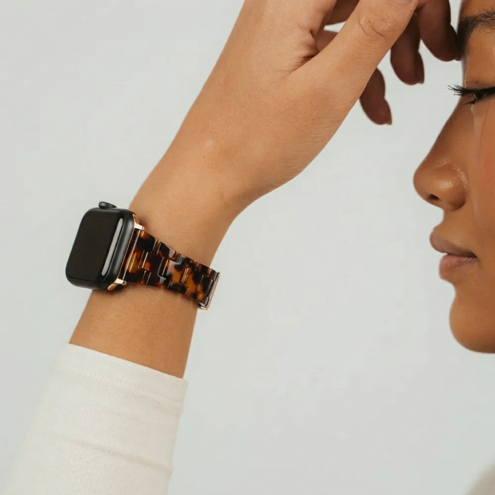 Person wearing a slim, tortoiseshell Apple Watch strap with gold hardware, hand raised near face.