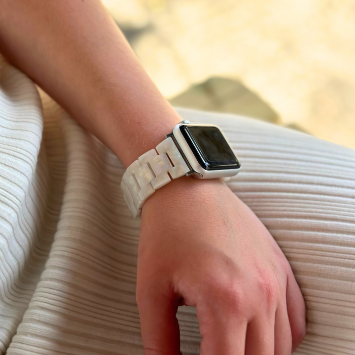 Close-up of a person's wrist adorned with the Luxe Iridescent Apple Watch Strap by Coconut Lane. Their hand rests on their lap, which is draped in white pleated fabric. The blurred background directs attention to the watch strap and hand.
