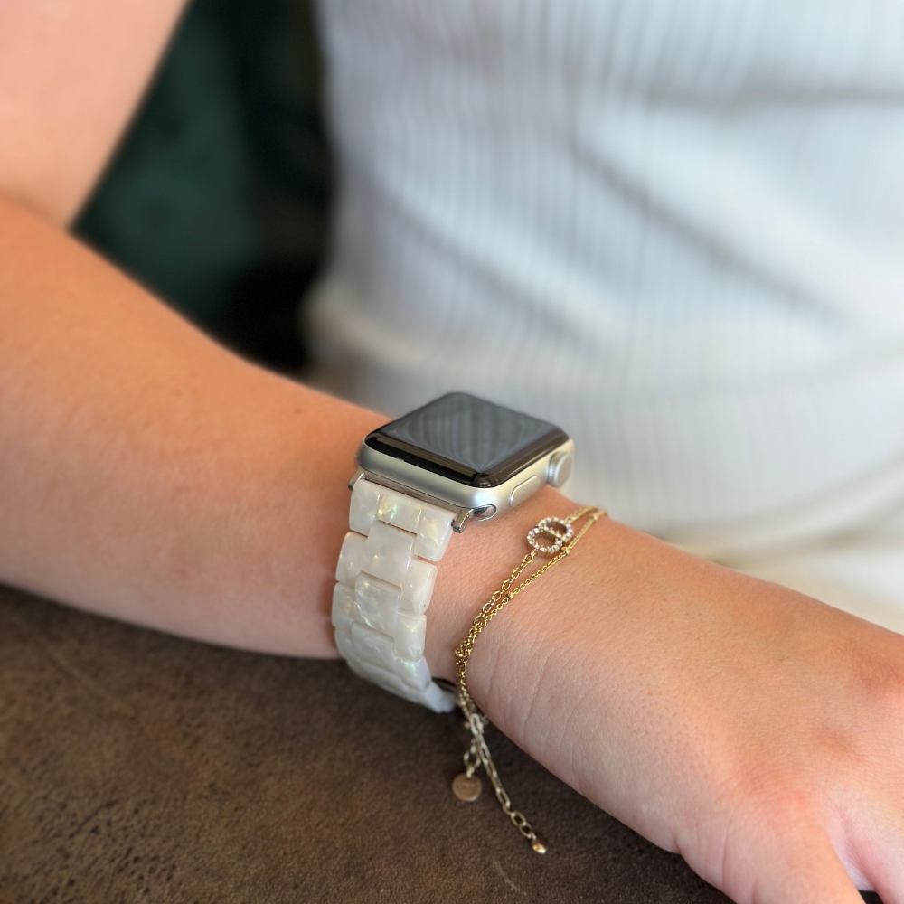 A close-up of an arm resting on a tabletop, adorned with the Luxe Iridescent Apple Watch Strap by Coconut Lane. Next to the watch is a delicate gold chain bracelet featuring a small charm. The person is wearing a white, sleeveless top.