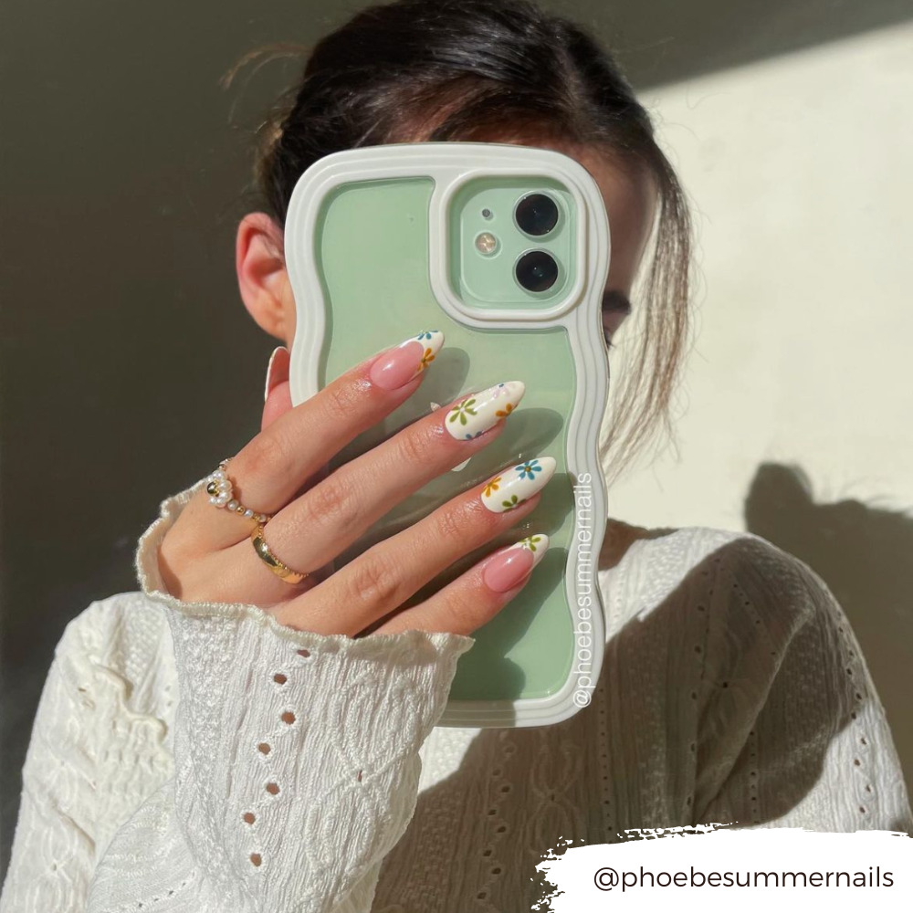 A person with dark hair, wearing a white knitted sweater, holds a green Coconut Lane Curvy Phone Case - Cream partially covering their face. Their nails are long with colorful daisy designs. The Instagram handle "@phoebesummernails" is visible in the image.