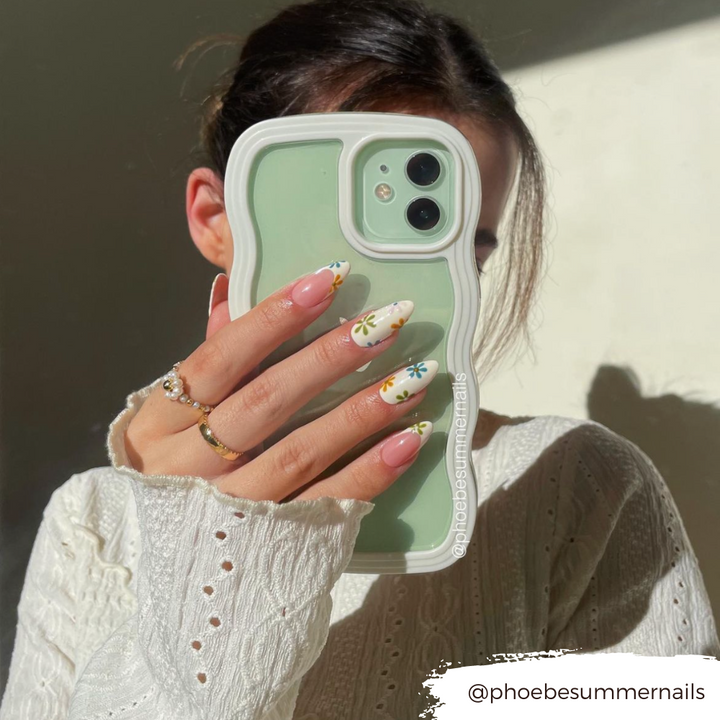 A person with dark hair, wearing a white knitted sweater, holds a green Coconut Lane Curvy Phone Case - Cream partially covering their face. Their nails are long with colorful daisy designs. The Instagram handle "@phoebesummernails" is visible in the image.