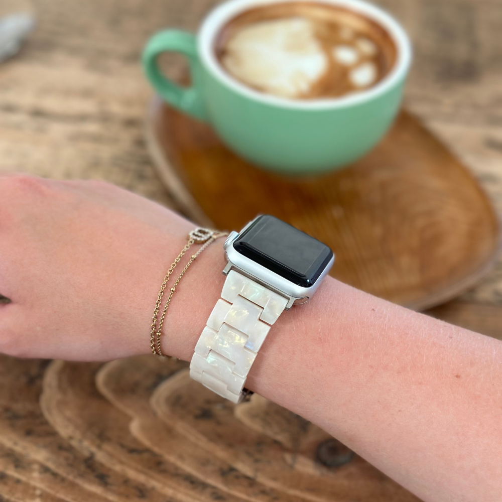A person wearing a Luxe Iridescent Apple Watch strap by Coconut Lane and a gold chain bracelet on their wrist, holding a wooden coffee stirrer. A cappuccino in a green mug sits on a wooden table in the background.
