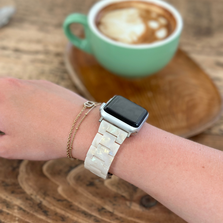 A person wearing a Luxe Iridescent Apple Watch strap by Coconut Lane and a gold chain bracelet on their wrist, holding a wooden coffee stirrer. A cappuccino in a green mug sits on a wooden table in the background.