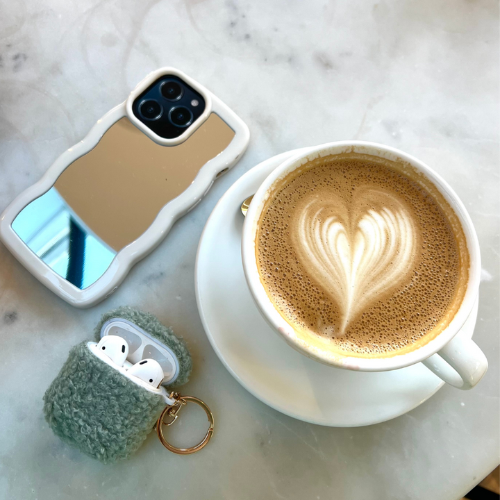 A cup of latte with heart-shaped latte art sits on a saucer atop a marble surface. Next to it are an iPhone adorned with the sleek and stylish Curvy Mirror Phone Case in cream by Coconut Lane, and a keychain holding a fuzzy gray AirPods case, with the AirPods partially visible.