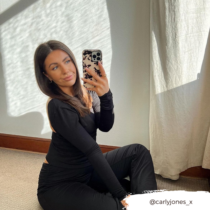A woman with long, dark hair sits on the floor in front of a light-colored wall, capturing a mirror selfie. She is dressed in a black long-sleeve top and black pants. Sunlight streams through a bright curtain, accentuating her Ivory Tort Phone Case from Coconut Lane on her iPhone 15. Her Instagram handle, @carlyjones_x, is displayed in the bottom right corner of the image.