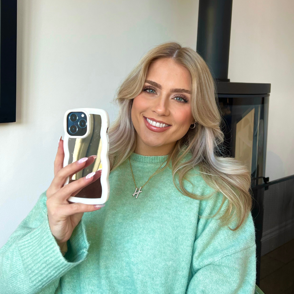 A person with long blonde hair, wearing a mint green sweater, smiles while holding up a smartphone encased in the sleek and stylish Curvy Mirror Phone Case - Cream by Coconut Lane. A necklace with a cross pendant hangs around their neck, and the background features a modern fireplace against a white wall.