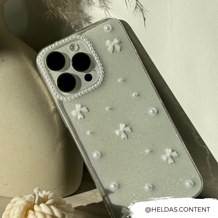 A close-up of a glittery, clear design Mini White Bows Phone Case for iPhone 15 by Coconut Lane, adorned with small white bows and pearls. The phone is leaning against a neutral-colored vase. The Instagram handle "@HELDAS.CONTENT" is displayed in the bottom-right corner of the image.