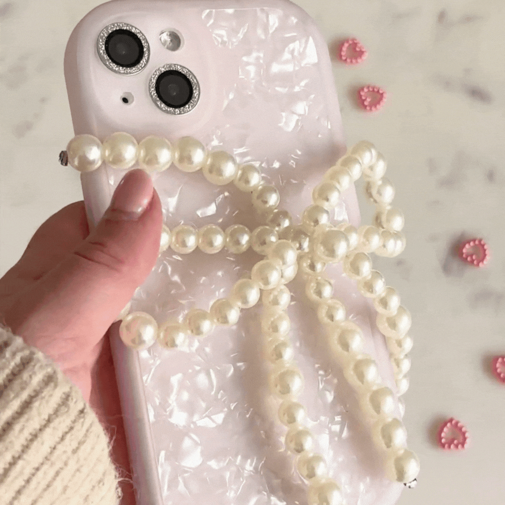 A close-up of a hand holding a phone with a pearlescent white case featuring Coconut Lane's Beaded Phone Strap - Pearl Bow. Small pink heart-shaped decorations are scattered on a marble surface in the background, and the person is wearing a beige knitted sleeve.
