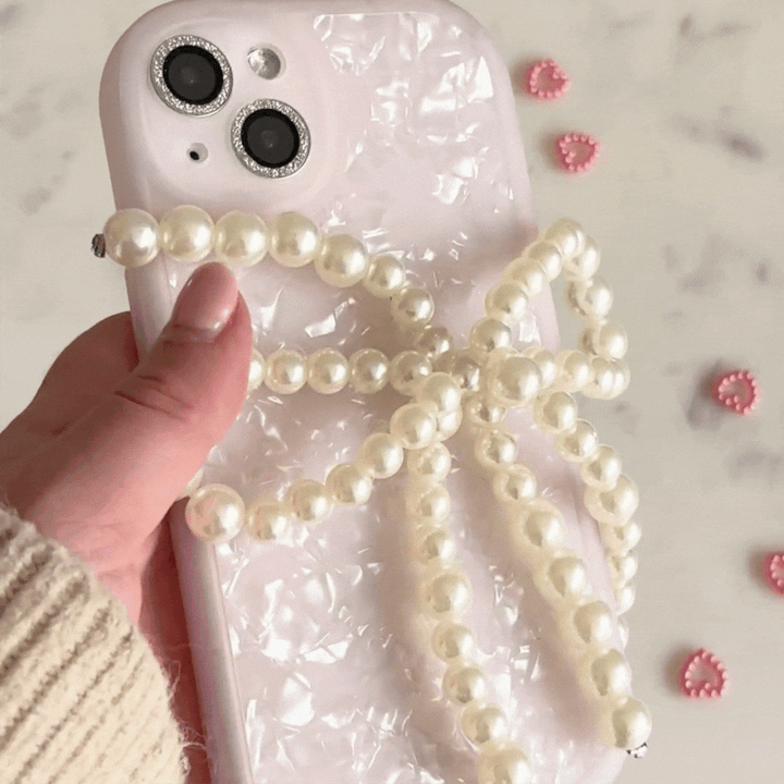 A close-up of a hand holding a phone with a pearlescent white case featuring Coconut Lane's Beaded Phone Strap - Pearl Bow. Small pink heart-shaped decorations are scattered on a marble surface in the background, and the person is wearing a beige knitted sleeve.
