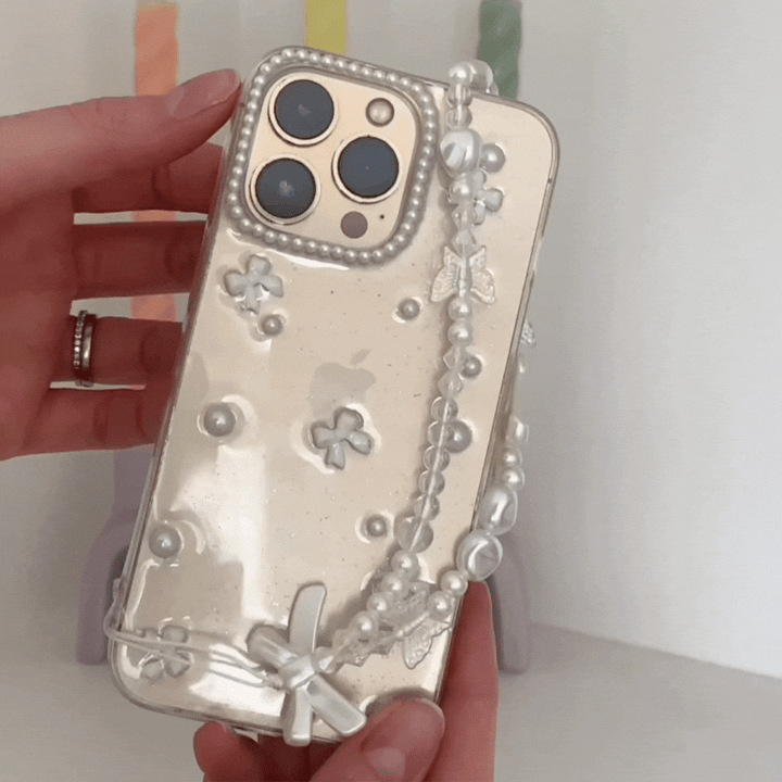 A hand is holding the Coconut Lane Mini White Bows Phone Case, adorned with a clear decorative design featuring pearl-like beads, small flower embellishments, and mini white bows. The background shows colorful, blurred objects.