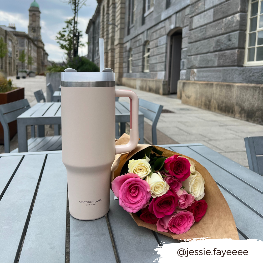 Marshmallow pink stainless steel tumbler by Coconut Lane on an outdoor table next to roses, with stone buildings in background.