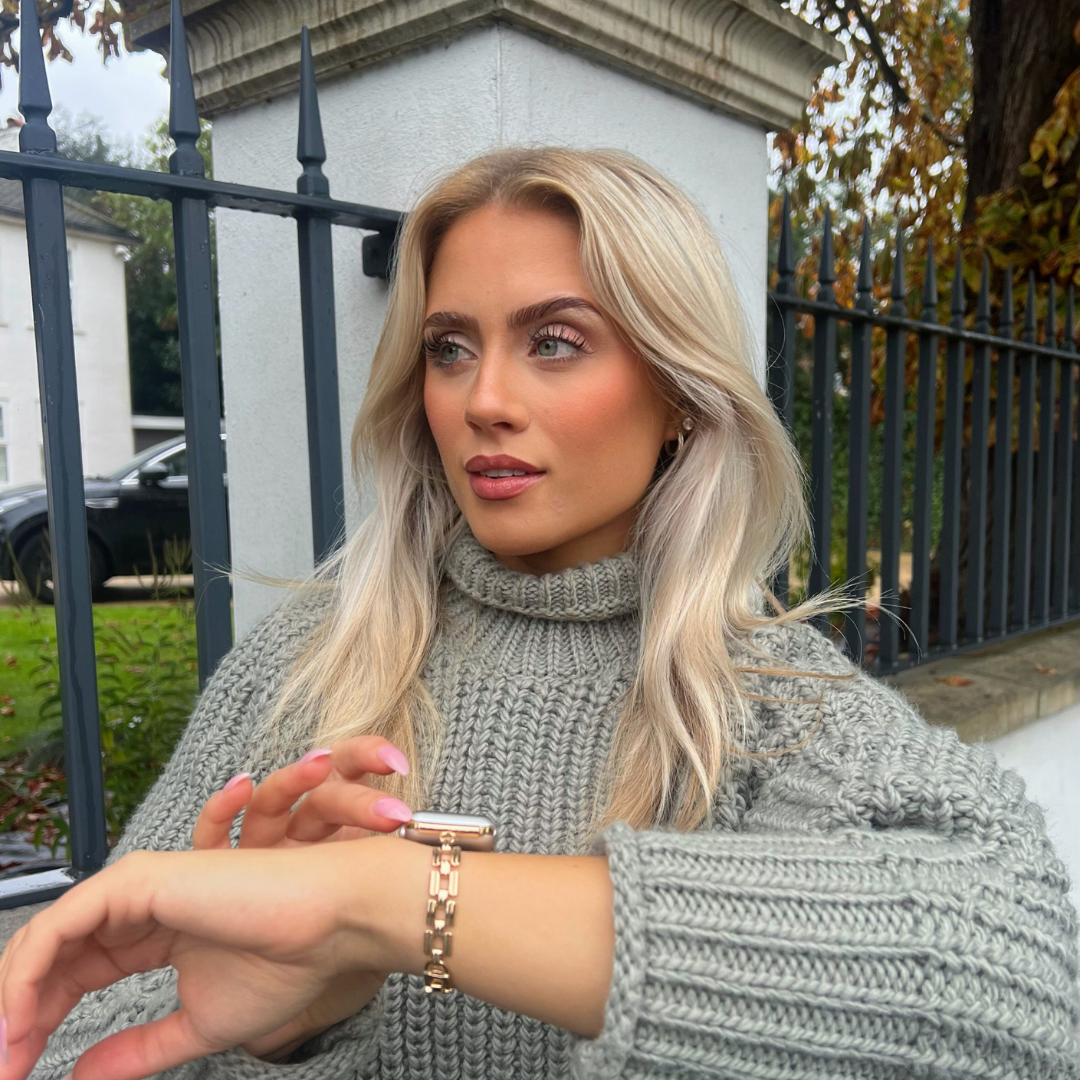 A woman with long blonde hair is standing outdoors in front of a black metal fence and a white building. She is wearing a gray turtleneck sweater and looking off to the side, while adjusting her Coconut Lane Square Gold Bracelet Apple Watch Strap with her other hand.