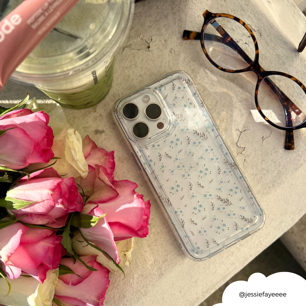 Clear phone case with blue floral pattern by Coconut Lane, shown on marble with pink roses, a drink, and glasses in sunlight.