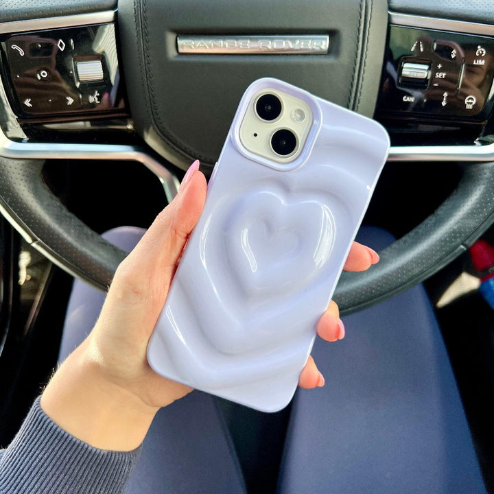 A person is holding a smartphone with Coconut Lane's Melting Heart Phone Case in shimmering lilac, featuring a raised heart design. The phone is positioned in front of a car steering wheel with the Range Rover brand logo visible. The person's nails are painted pink, and they are wearing a long-sleeve top.