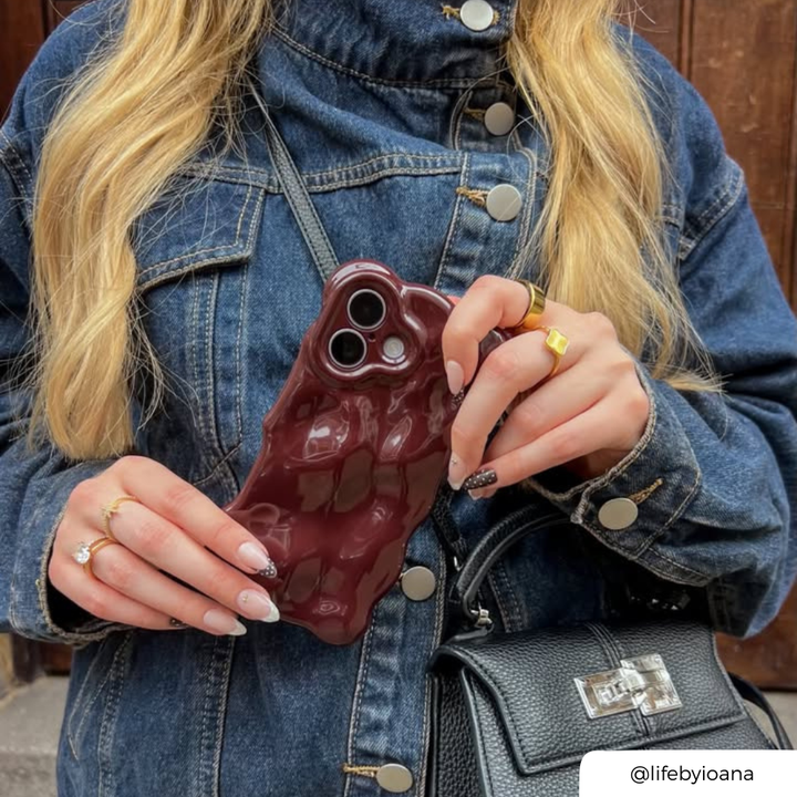 A woman with long blonde hair, wearing a dark blue denim jacket in front of a brown door holds an iphone up to her ear. The phone is in a 3d bubble burgundy phone case.