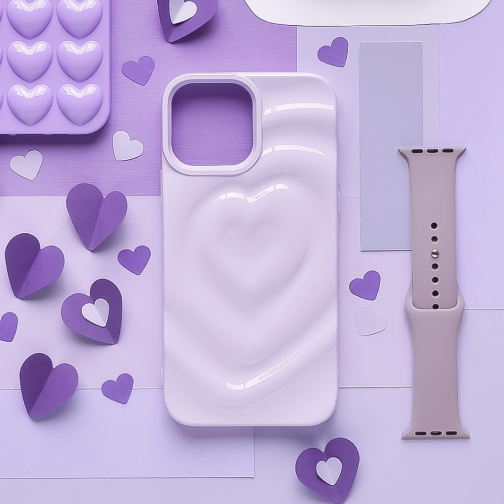 A textured Melting Heart Phone Case in pastel lilac from Coconut Lane is placed on a table surrounded by shimmering lilac and heart-shaped objects. There is also a tan-colored smartwatch band and a lavender silicone mold with heart shapes. The background consists of various purple-toned materials.