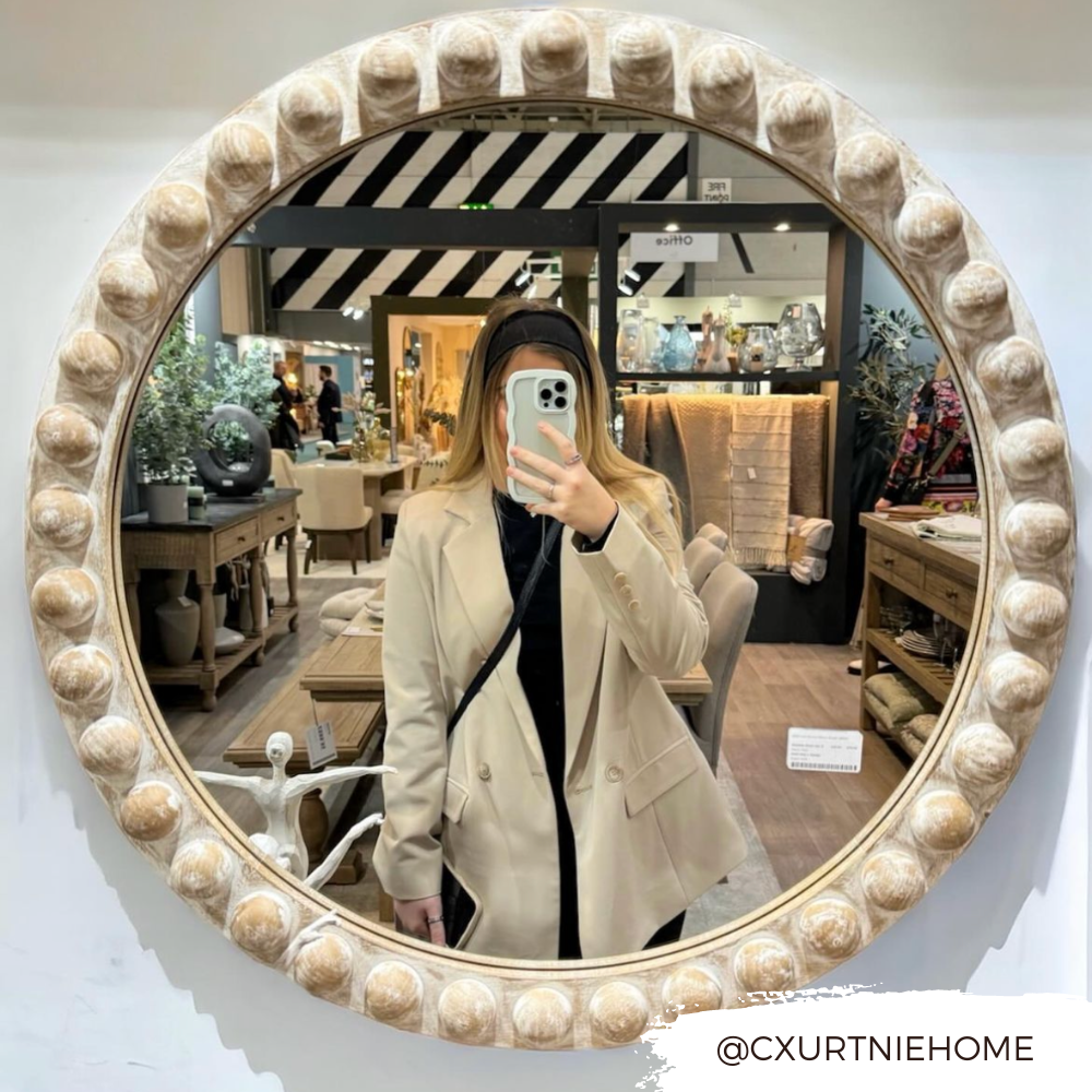 A person wearing a beige blazer and black top takes a mirror selfie in a home decor store. Various items like furniture and decorative pieces are displayed in the background. The mirror has a round, ornate frame with a beaded design. They're holding the Curvy Phone Case - Cream by Coconut Lane, which is highly protective for their iPhone 15. Text in the corner reads "@CXURTNIEHOME.