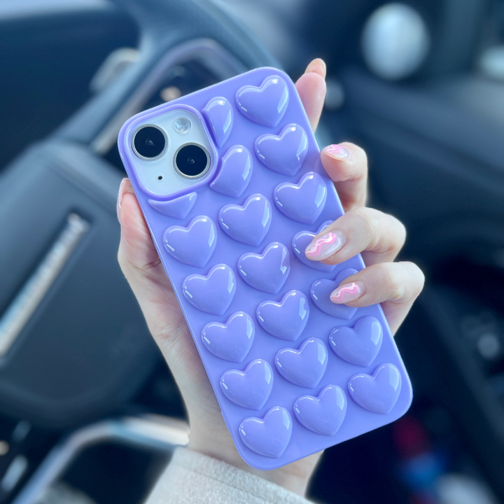 A hand with long, light pink manicured nails holds a Coconut Lane 3D Hearts Phone Case in Deep Lilac, adorned with raised heart-shaped designs. In the background, a blurred car interior featuring a steering wheel and controls can be seen.