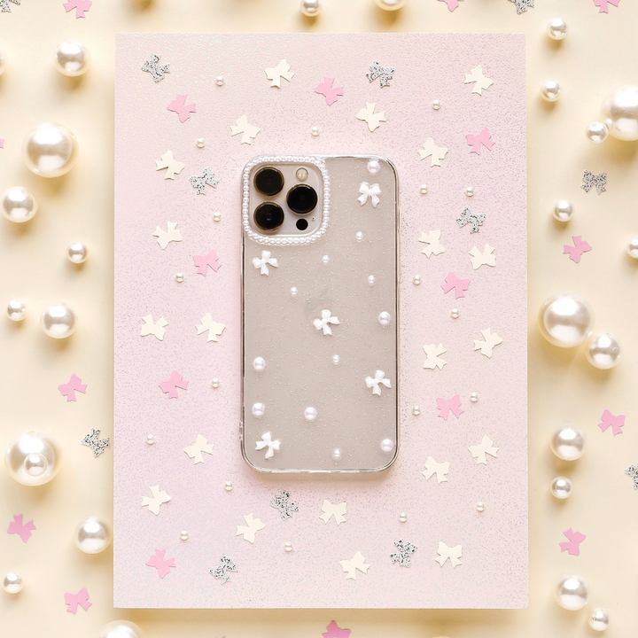 A smartphone with a Coconut Lane Mini White Bows Phone Case featuring a transparent floral and pearl design is placed on pink paper, adorned with small shiny decorations. The backdrop showcases a pastel color scheme with scattered pearls, pink stars, and glittery silver leaf shapes.