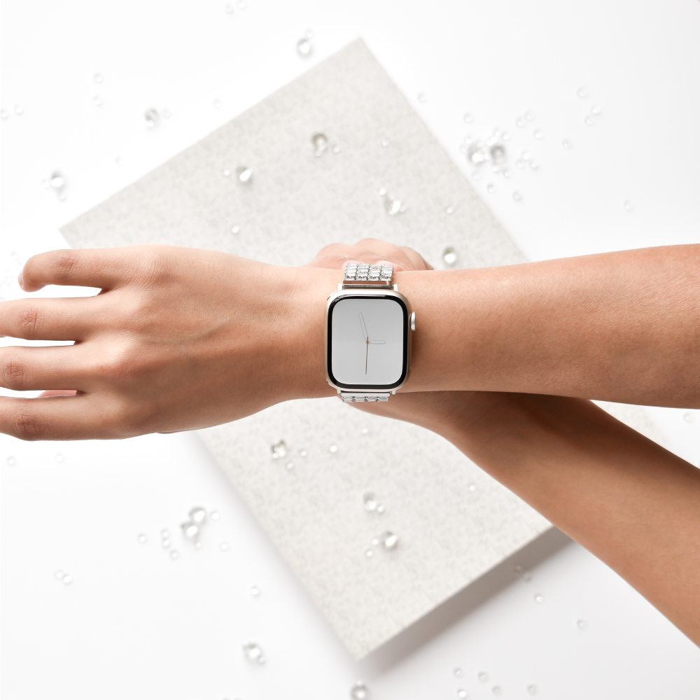 A close-up of an arm wearing a stylish smartwatch with the Sparkle Super Glam Apple Watch Strap by Coconut Lane, featuring an adjustable stainless steel strap and a square face. The background displays a textured, light grey surface adorned with scattered small water droplets. The watch screen is blank, displaying no content.