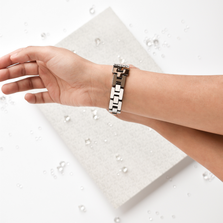A close-up of a person's wrist wearing the stylish Sparkle Super Glam Apple Watch Strap by Coconut Lane, featuring a geometric link design and an adjustable strap. The background showcases a white surface adorned with scattered pearly beads and a rectangular textured white object, creating an overall minimalistic and elegant scene.