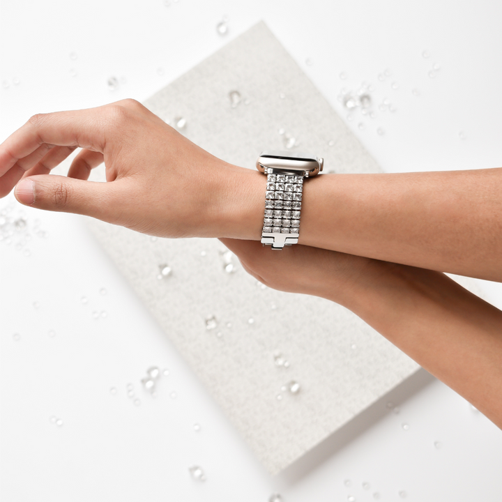 A close-up of an individual wearing the Sparkle Super Glam Apple Watch Strap by Coconut Lane. The person's wrist is extended over a gray textured surface, with separated small glass or crystal beads dotting the white background.