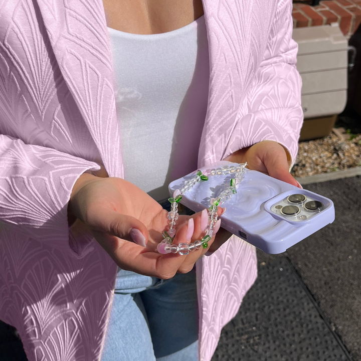 A person wearing a textured pink blazer and white top holds a smartphone adorned with a shimmering lilac beaded charm. The phone, featuring the light-colored, Melting Heart Phone Case - Lilac by Coconut Lane, is held against the backdrop of a sunny day outdoors.
