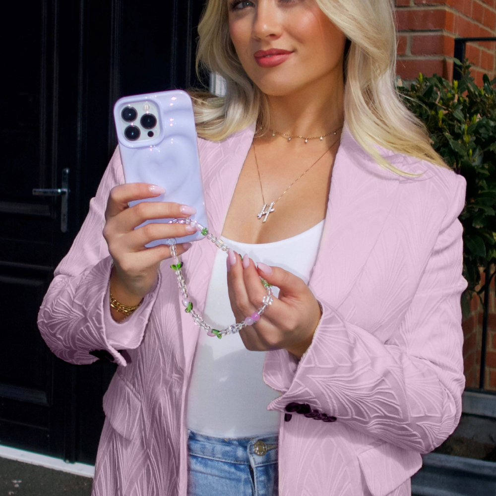 A blonde woman stands outdoors holding a smartphone encased in the Melting Heart Phone Case - Lilac by Coconut Lane, complete with a decorative beaded strap. She wears a shimmering lilac blazer over a white top and light blue jeans. A necklace with a cross pendant is visible around her neck. Brick and greenery form the backdrop.

