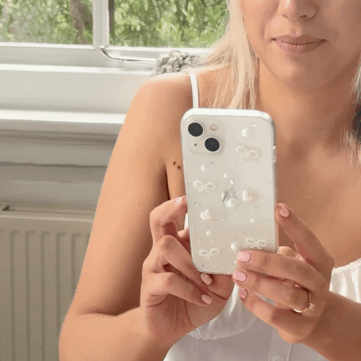 A person with long blonde hair is holding a Coconut Lane Coquette Dreams Phone Case in their right hand. They sit near a window with green foliage visible outside, wearing a white sleeveless top and a ring on their left hand.