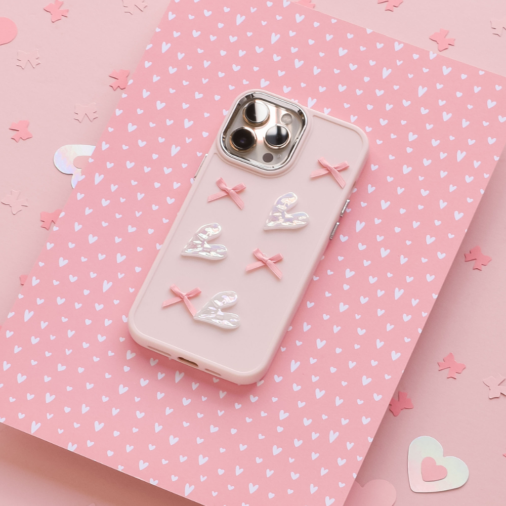 A Coconut Lane Pearly Hearts Phone Case - Pink, designed for the iPhone 15 and adorned with four white hearts and three pink bows, lies flat against a pink background featuring white heart patterns and scattered heart-shaped confetti. The phone's rear cameras are visible through cutouts in the case.