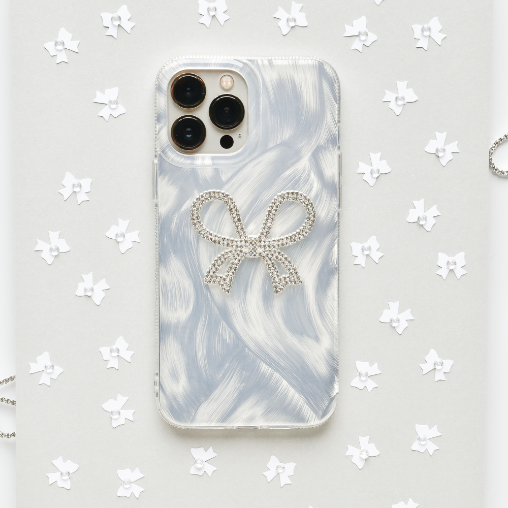 A Coconut Lane Diamante Bow Phone Case, featuring a marbled light grey design and adorned with a diamante bow decoration, is surrounded by small white paper stars on a light grey background.