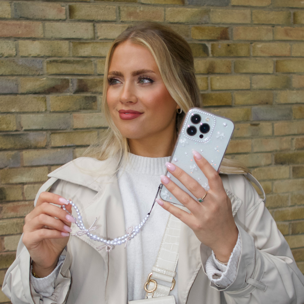 A woman with long blonde hair is holding a smartphone featuring the Mini White Bows Phone Case from Coconut Lane, along with a beaded strap. She is dressed in a light-colored coat and sweater, set against a brick wall backdrop. Her nails are painted light pink, and she seems to be gazing slightly off-camera.
