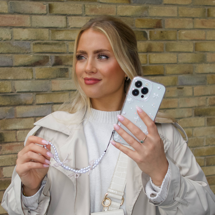 A woman with long blonde hair is holding a smartphone featuring the Mini White Bows Phone Case from Coconut Lane, along with a beaded strap. She is dressed in a light-colored coat and sweater, set against a brick wall backdrop. Her nails are painted light pink, and she seems to be gazing slightly off-camera.