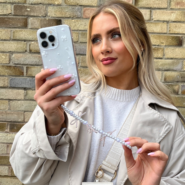 A woman with long blonde hair holds up her smartphone, encased in a Coconut Lane Mini White Bows Phone Case, to take a selfie. She is wearing a light tan coat over a white top. Her phone’s beaded strap complements the clear design of the iPhone 15 case. The background features a brick wall.