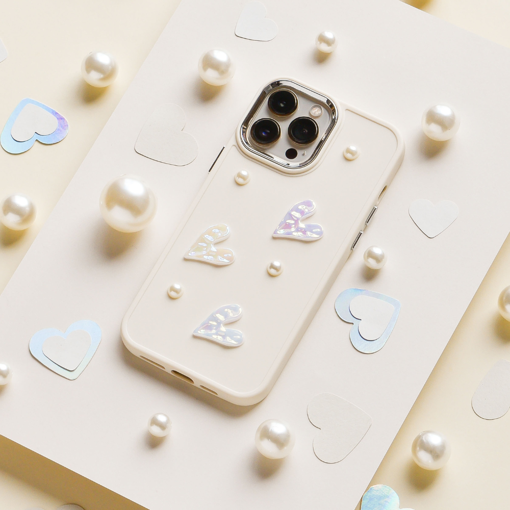 The Pearly Hearts Phone Case - Nude by Coconut Lane, adorned with three pearlescent hearts and small pearls, is showcased amidst scattered white and iridescent heart cutouts and spherical pearls on a light-colored background, providing an extra layer of protection.