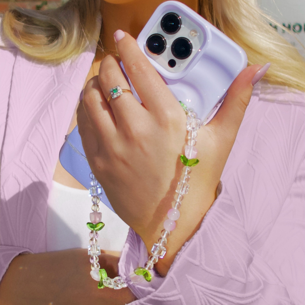 A person in a shimmering lilac outfit holds a light purple smartphone encased in the Melting Heart Phone Case - Lilac by Coconut Lane, featuring a textured design and an attached beaded strap adorned with clear, pink, and green leaf-shaped beads. They also wear a square-cut gemstone ring and have manicured nails painted light pink.