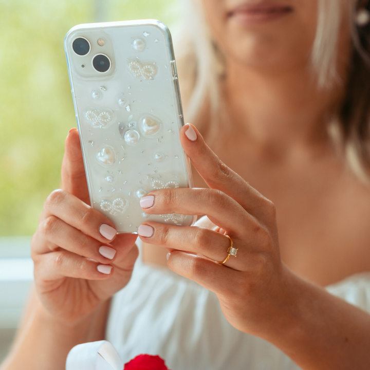 A person with light pink manicured nails is holding an iPhone 15 with a Coquette Dreams Phone Case by Coconut Lane, adorned with white pearls. They are wearing a white top, and a delicate gold ring with a clear gemstone is visible on their left hand. A blurred green background provides a serene backdrop.