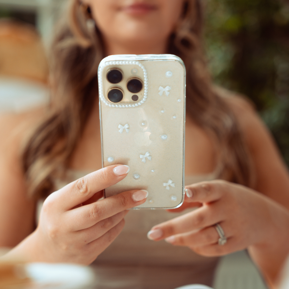 A person with wavy hair holds a smartphone encased in a Coconut Lane Mini White Bows Phone Case, featuring a decorative design adorned with small white flowers and pearls. The person wears a ring on their left ring finger, while the background remains blurred with green foliage and warm-toned elements.