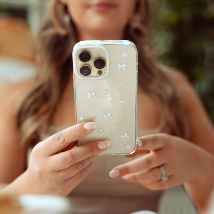 A person with wavy hair holds a smartphone encased in a Coconut Lane Mini White Bows Phone Case, featuring a decorative design adorned with small white flowers and pearls. The person wears a ring on their left ring finger, while the background remains blurred with green foliage and warm-toned elements.