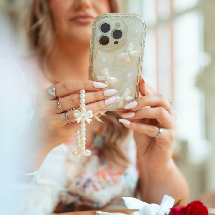 A woman with manicured nails holds the Pearls & Bows Phone Case by Coconut Lane, adorned with pearls and bows. She wears rings and a bracelet that match the phone's decoration. The background is softly blurred, focusing on her hands and the protective phone case.