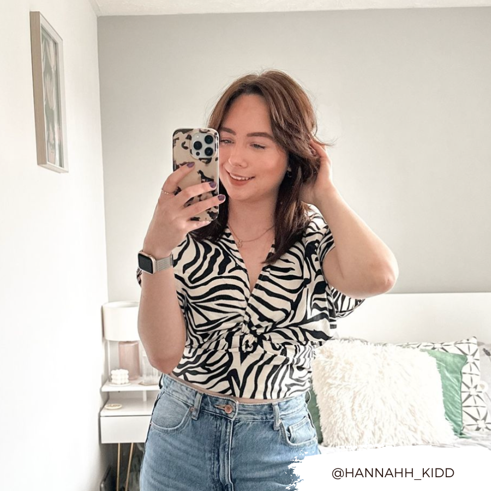 In a bright bedroom, a woman smiles at her phone while taking a mirror selfie. She is wearing a black and white zebra print top and jeans, holding an Ivory Tort Phone Case by Coconut Lane. The room showcases a bed adorned with white and green pillows, a nightstand, and minimalist decor. The handle @HANNAHH_KIDD is visible in the corner.