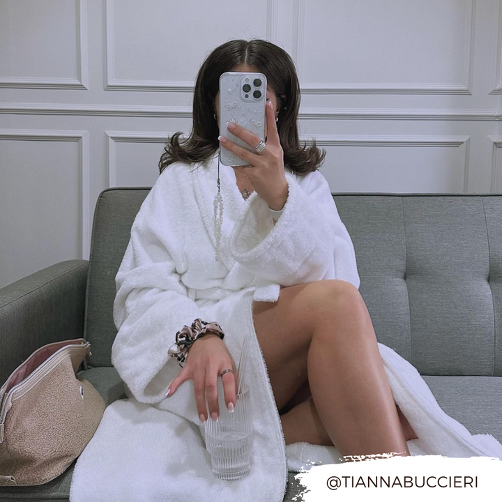 A person with shoulder-length hair sits on a gray couch wearing a white robe. They are holding an iPhone15 with a Coquette Dreams Phone Case by Coconut Lane up to their face, and a glass of water in their other hand. A beige bag and some accessories are next to them on the couch. The Instagram handle @TIANNABUCCIERI is displayed in the lower right corner.