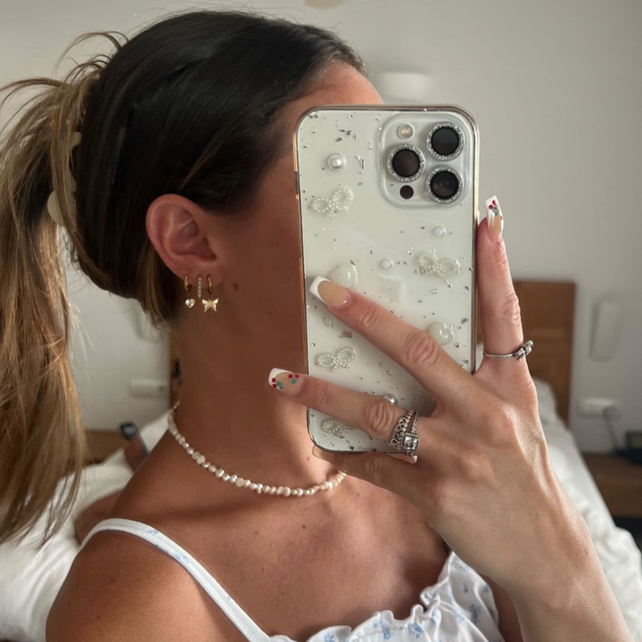 A woman with a ponytail takes a mirror selfie, partially covering her face with her Coconut Lane Coquette Dreams Phone Case. She sports butterfly-shaped earrings, a pearl necklace, and flaunts a ring-adorned hand with manicured nails painted in a floral design. Her iPhone15 case stands out, adding an extra protective charm to the bedroom snapshot.